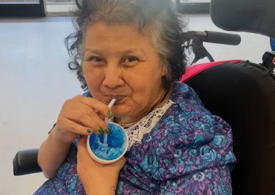 A smiling female resident in a wheelchair enjoying a blue shaved ice drink through a straw.